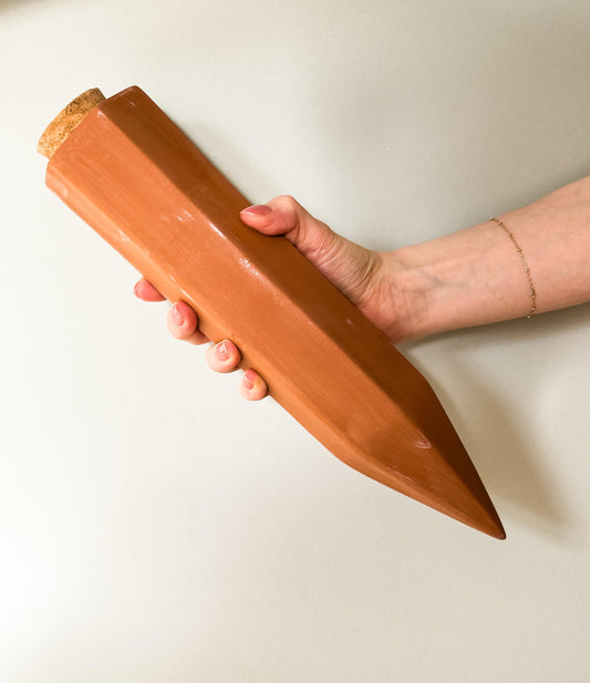 A terracotta automatic watering spike held in a person's hand, with a cork stopper at one end.